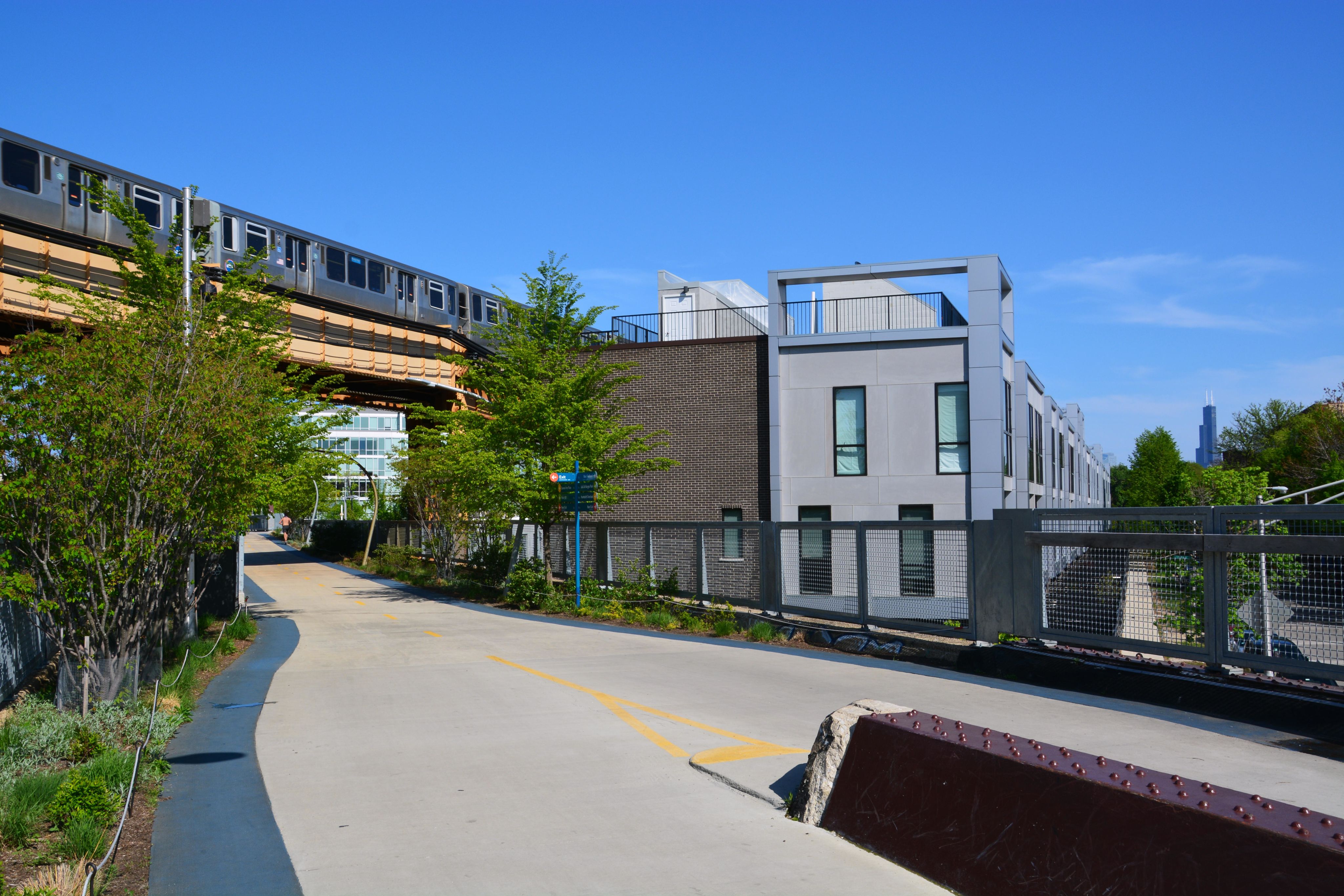 A luxury condominimum building with dark grey brick and light grey window framings is the focus of this image. The building is mere feet from the 606 trail, which is in the foreground of the image. A blue line train runs over the trail on a blue sky day. 